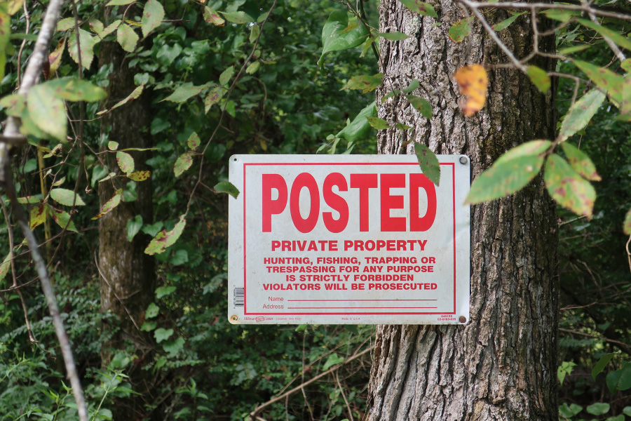 A sign attached to a tree in the woods. It reads, "Posted: Private property. Hunting, fishing, trapping, or trespassing for any purpose is strictly forbidden. Violators will be prosecuted."